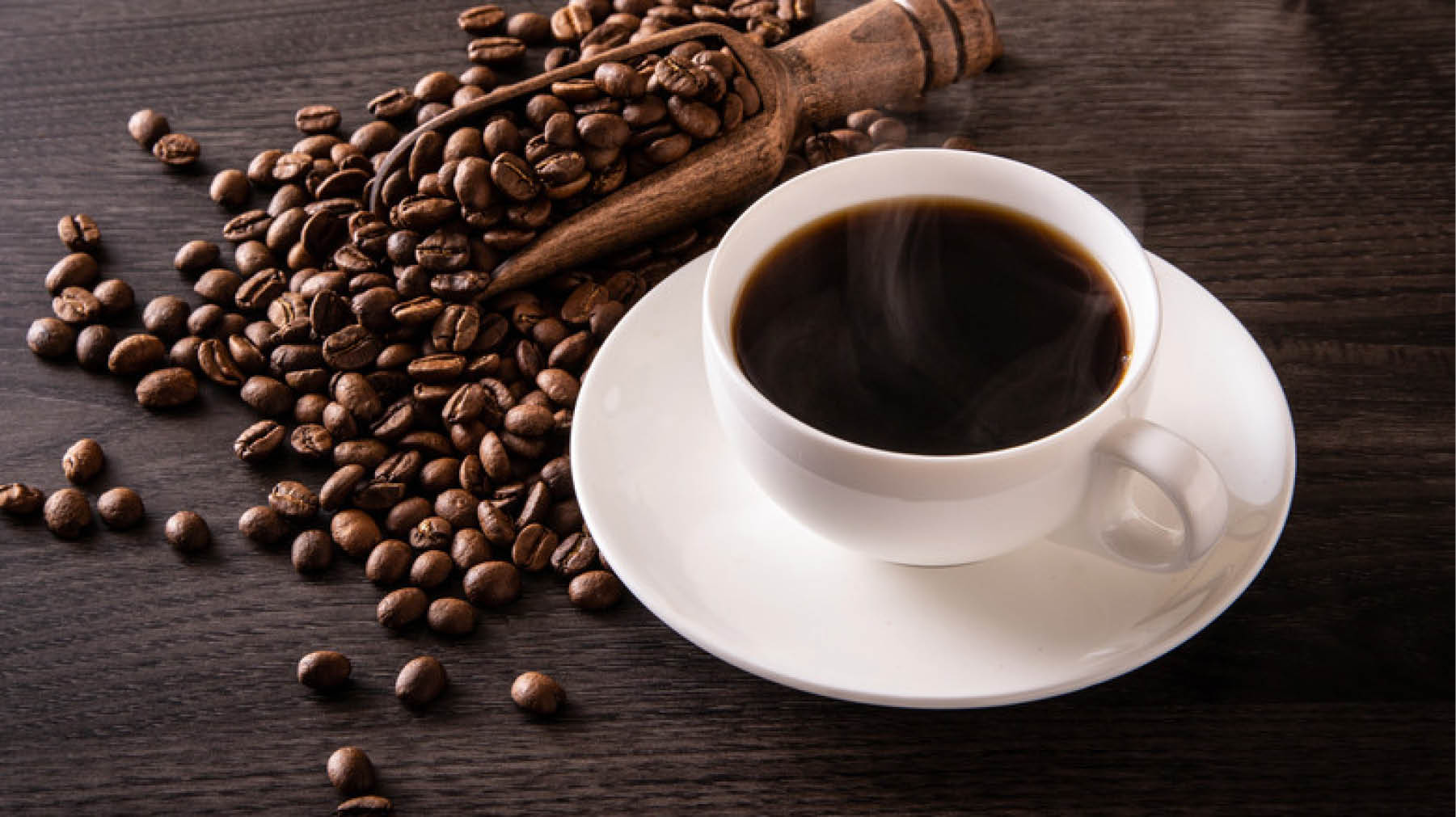 A steaming cup of black coffee sits on a white saucer next to a wooden scoop filled with coffee beans scattered on a dark wooden surface. Description generated by AI